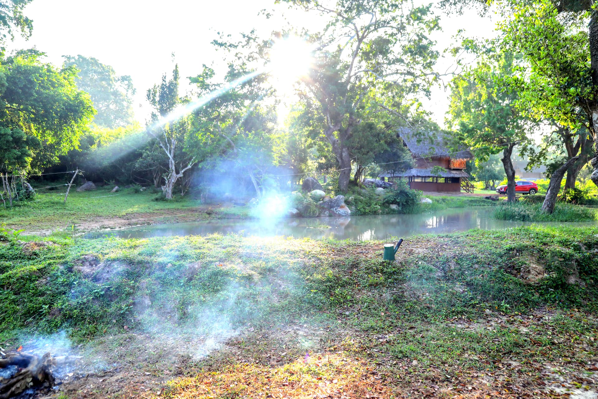 A sunlit view of the traditional thatched-roof cabana beside a tranquil pond, with a campfire area set for an evening gather.